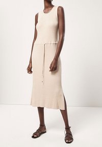 Robe midi beige côtelée avec encolure ronde, sans manches, taille ajustable avec cordon, et fentes latérales. Portée avec des sandales noires à brides.