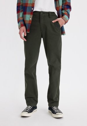 Pantalons vert foncé à coupe droite, fabriqués en coton, dotés de poches avant et portés avec des baskets noires et une chemise à carreaux.