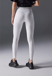 Valkoiset korkeavyötäröiset legginsit, joissa on sileä pinta ja tiukka muotoilu, yhdistettynä mustiin urheilukenkihin, nähtynä takaa.