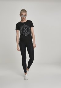 Sort t-shirt med et hvidt geometrisk Linkin Park grafik, parret med sorte leggins og hvide sneakers. Glat stof, tætsiddende design.