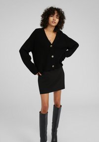 Schwarze gerippte Strickjacke mit V-Ausschnitt und Knopfverschluss, kombiniert mit einem taillierten schwarzen Rock. Das Model trägt graue kniehohe Stiefel.
