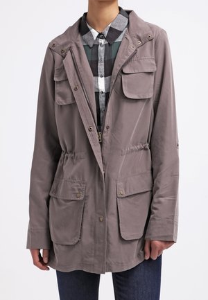 Personne portant une veste à capuche taupe avec une fermeture éclair frontale et des poches à pression sur une chemise à carreaux noire, blanche et verte et un jean bleu foncé.