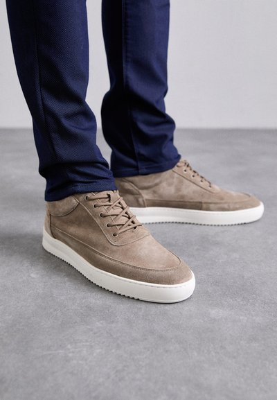 Zamszowe buty casual w jasnobrązowym kolorze z białą gumową podeszwą. Posiadają okrągłe sznurówki oraz teksturalne detale. Noszone z granatowymi spodniami.