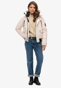 Jasnoróżowa kurtka puchowa z czarnym zamkiem i trzema kieszeniami, noszona na beżowym swetrze z dzianiny; w zestawie z podwijanymi niebieskimi dżinsami i czarnymi butami.