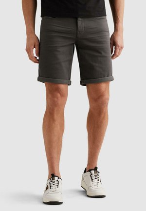 Muž v tmavosivých kraťasoch vyhrnutých nad kolená, čiernom tričku a bielych teniskách stojaci na jednoduchom svetlom pozadí.