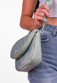 Šedá, měkká látková taška zaobleného tvaru, se zipovým uzávěrem, nastavitelným popruhem a malým vyšitým logem vpředu.