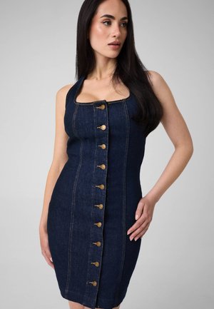 Donna con lunghi capelli scuri che indossa un vestito di jeans blu scuro senza maniche con bottoni dorati sul davanti, posando su uno sfondo grigio chiaro uniforme.