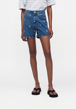 Person iført højtaljede blå denimshorts med små hvide blomsterbroderier, kombineret med sorte platåsandaler og let stribet top.