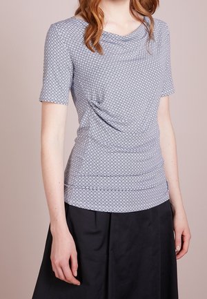 Kobieta ubrana w dopasowaną bluzkę z krótkim rękawem o wzorze z dekoltem w kształcie opadającej tkaniny i czarną spódnicę, stojąca na jednolitym tle.