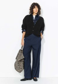 Cardigan noir en maille avec un col en V, associé à une chemise en denim et un pantalon large à rayures navy. Les accessoires comprennent un sac en imprimé léopard et des mocassins noirs.