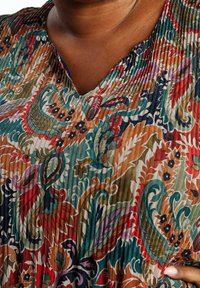 Haut en tissu plissé multicolore avec un col en V, présentant des motifs floraux et paisley complexes dans des tons verts, rouges, bleus et orange.