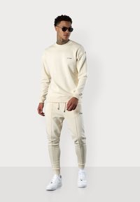 Beige Sweatshirt mit langen Ärmeln und Rundhalsausschnitt, kombiniert mit passenden, schmal geschnittenen Jogginghosen, getragen mit weißen Sneakers und schwarzen Sonnenbrillen.