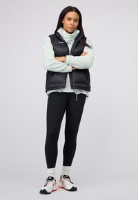 Schwarze Pufferweste über einem hellgrünen Fleece-Oberteil, kombiniert mit schwarzen Leggings und weißen Sportschuhen mit orangefarbenen Akzenten.