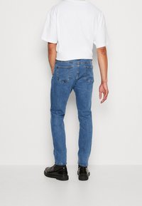 Blue denim jeans with a slim fit, five pockets, and a clean design. Paired with a plain white t-shirt and black shoes.