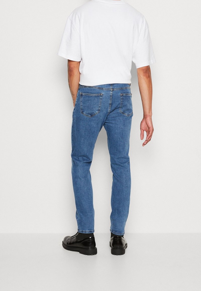 Blue denim jeans with a slim fit, five pockets, and a clean design. Paired with a plain white t-shirt and black shoes.