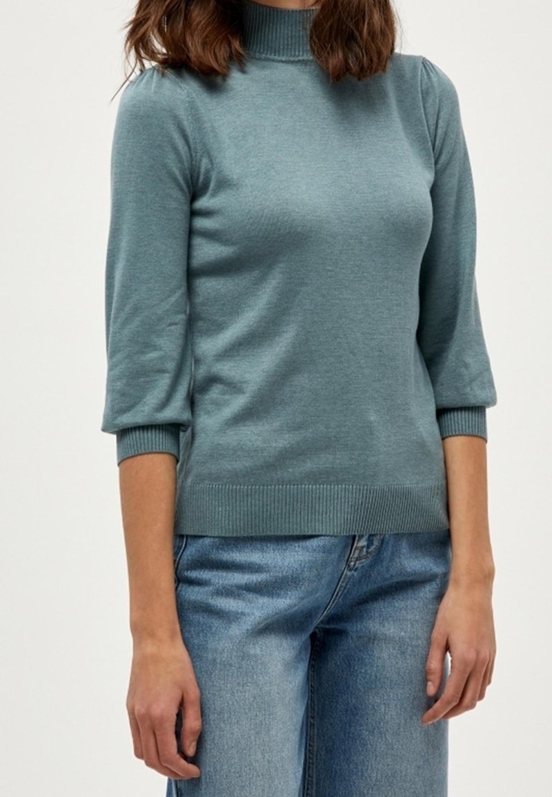Femme portant un pull à col roulé côtelé bleu sarcelle avec des manches trois-quarts et un jean bleu sur un fond uni.