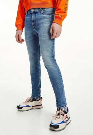 Homme portant un jean skinny bleu délavé, des baskets blanches et bleues avec des lacets roses, et un sweat-shirt orange avec un texte sur la manche.