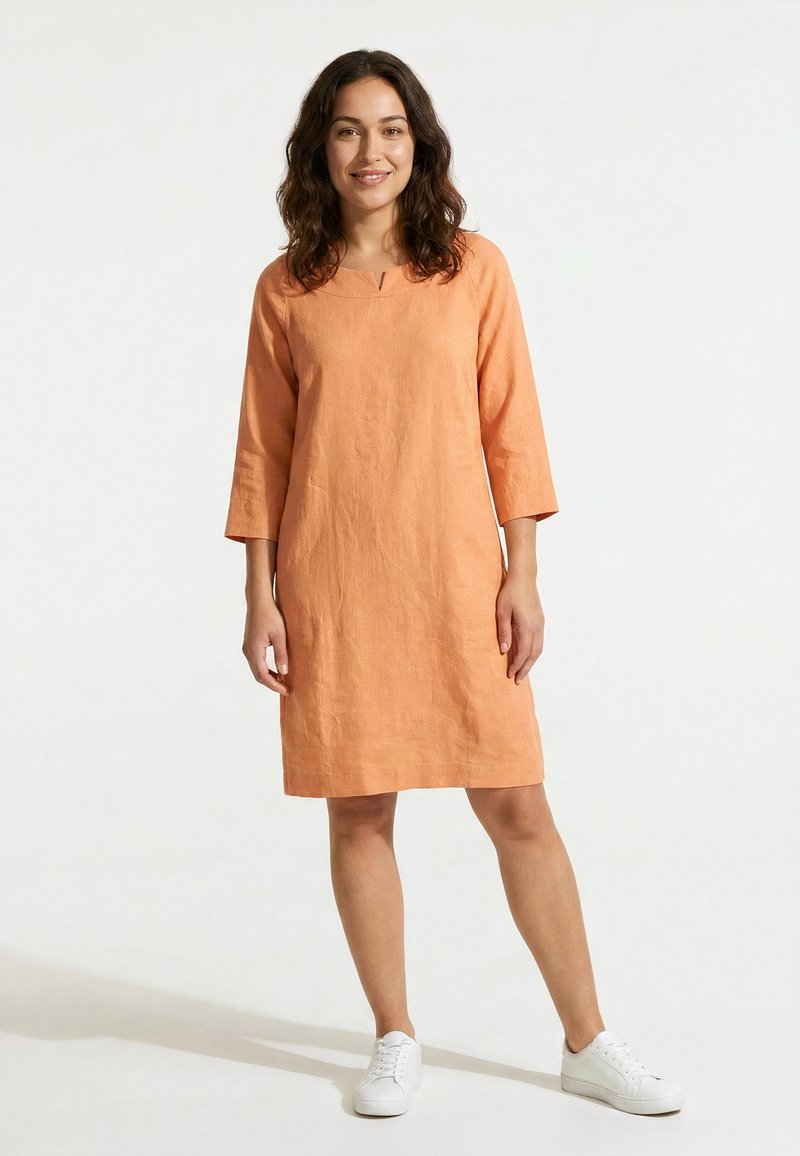 Femme aux cheveux bouclés foncés portant une robe orange longueur genoux à manches longues et des baskets blanches, debout sur un fond blanc.