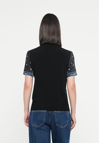Black sleeveless top with a textured back and patterned short sleeves featuring blue floral accents and white dots. Denim pants visible.