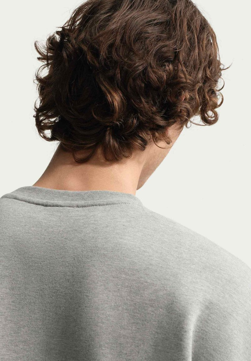 Personne aux cheveux bruns bouclés vue de dos portant un t-shirt gris clair à col rond sur un fond uni.