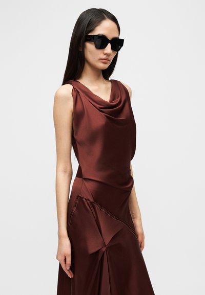Καφέ σατέν φόρεμα με ντραπέ λαιμόκοψη και μοναδικές λεπτομέρειες πτυχωμάτων, συμπληρωμένο με μαύρα γυαλιά ηλίου με γεωμετρικό σκελετό.