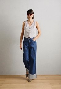 Vest gri deschis în carouri cu butoane, asortat cu blugi evazați din denim albastru închis cu manșete pliate. Femeie purtând ochelari de soare roz.