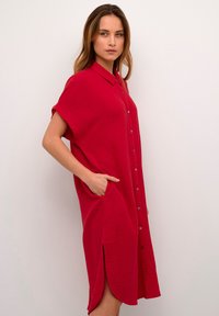Abito camicia rosso con bottoni, maniche corte, dotato di tasche laterali e tessuto con texture, che arriva appena sopra il ginocchio con un orlo leggermente curvo.