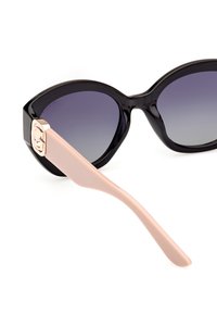 Gafas de sol estilo gato negro con lentes gradientes morados y brazos en color rosa claro que presentan un acento de logo plateado en la bisagra.