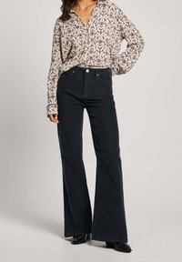 Camisa de manga larga floreada en crema con patrones rojos y azules, combinada con pantalones de campana negros de cintura alta y botas negras hasta el tobillo.