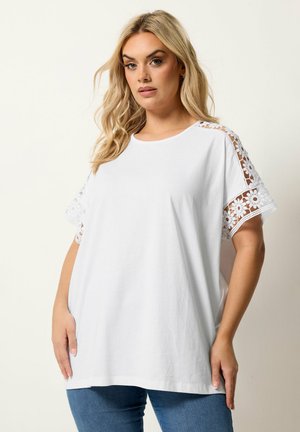 Blondynka ubrana w luźną białą bluzkę z kwiatowymi koronkowymi wycięciami na ramionach i rękawach, w połączeniu z niebieskimi dżinsami.