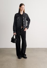 Musta farkkutakki, jossa on niittikoristeita, yhdistetty mustaan yläosaan ja leveälahkeisiin housuihin, pitäen pientä mustaa käsilaukkua. Sileä pinta.