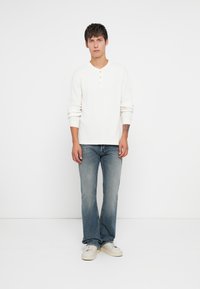 Chemise henley blanche à manches longues avec trois boutons, associée à un jean bleu clair et des baskets blanches, sur fond uni.