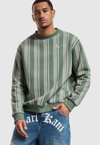 Felpa verde con strisce bianche verticali, colletto tondo e un piccolo logo ricamato sul petto. Indossata con jeans blu logori.