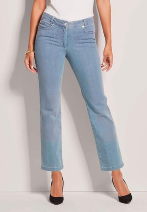 Vrouw die lichtblauwe jeans met rechte pijpen en zwarte hakken draagt, staand tegen een effen lichte achtergrond met ontspannen handen.