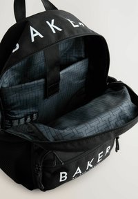Schwarzer Rucksack mit weißem "BAKER"-Schriftzug, ausgestattet mit einem strukturierten schwarzen Innenraum mit geometrischem Muster und einem Reißverschlussfach.