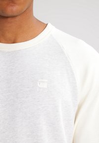 Světle šedý sweatshirt s raglánovými rukávy, měkkou texturou a diskrétním logem značky v horním levém rohu.