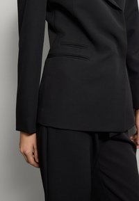 Primer plano de una persona que lleva un traje negro a medida con una chaqueta ajustada y pantalones a juego, contra un fondo simple.