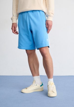 Persoon draagt lichtblauwe Adidas-shorts, beige sweatshirt, witte sokken en gebroken witte sneakers, staand op een blauwe vloer tegen een witte muur.