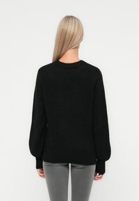 Pull en tricot noir, fuzzy, avec un col rond et des manches longues bouffantes. Ourlet et poignets côtelés ; texture douce. Porté par-dessus un jean gris ajusté.