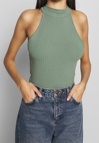 Bodysuit verde acanalado con cuello alto y hombros descubiertos, combinado con jeans de denim azul de cintura alta. La textura es suave y el ajuste es ceñido.