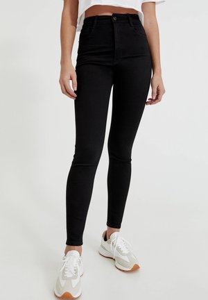 Woman wearing high-waisted black skinny jeans and white sneakers with beige accents, standing against a plain light background.