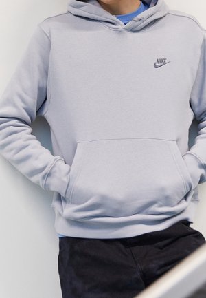 Osoba nosząca jasnoszary kaptur Nike z rękami w przedniej kieszeni i czarne spodnie, pod spodem widoczna niebieska koszula.