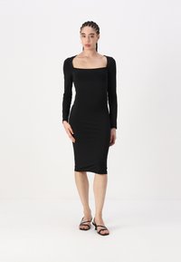 Ilga suknelė juoda, prigludusi prie kūno, su kvadratiniu iškirpimu, pagaminta iš lygaus audinio, iki kelių, su šoninėmis raukšlėmis. Suporuota su juodais strapiniais sandalais.