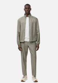 Svetlozelená bunda s vysokým golierom, spárovaná s ladiacimi nohavicami a bielou košeľou. Na dokončenie outfitu sú obuvou pohodlné topánky. Hladká textúra.