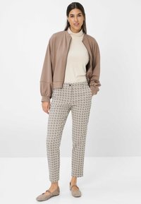 Hellbraune Bomberjacke über einem cremefarbenen Rollkragenpullover, gemusterte Slim-Fit-Hose in Beige mit geometrischen Designs und graue Ballerinas.