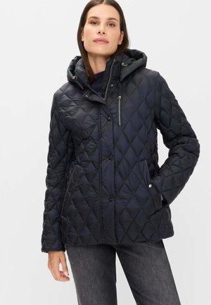 Frau trägt eine schwarze gesteppte Jacke mit Kapuze, Reißverschluss und Knopfleiste sowie Taschen, kombiniert mit dunkelgrauen Jeans.