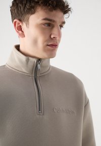 Felpa con zip di colore beige, caratterizzata da un colletto alto, dettagli delle cuciture discreti e logo ricamato 'Calvin Klein' sul petto. Texture liscia.