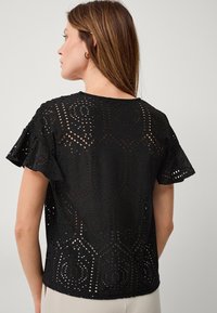 Schwarzes Spitzenoberteil mit hexagonalem Muster, kurzen Rüschenärmeln und gewelltem Saum, das filigrane Ausschnittdetails zeigt.