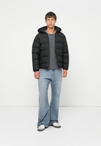 Jeune homme se tenant debout portant une doudoune noire, un pull en maille gris, un jean large bleu clair et des baskets blanches sur fond blanc.