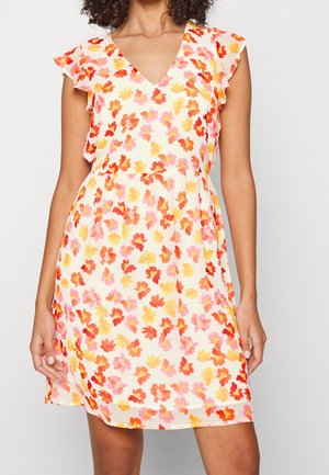 Donna che indossa un vestito senza maniche con scollatura a V, decorato con un motivo floreale rosso, arancione e rosa su sfondo bianco, in piedi con le mani rilassate.